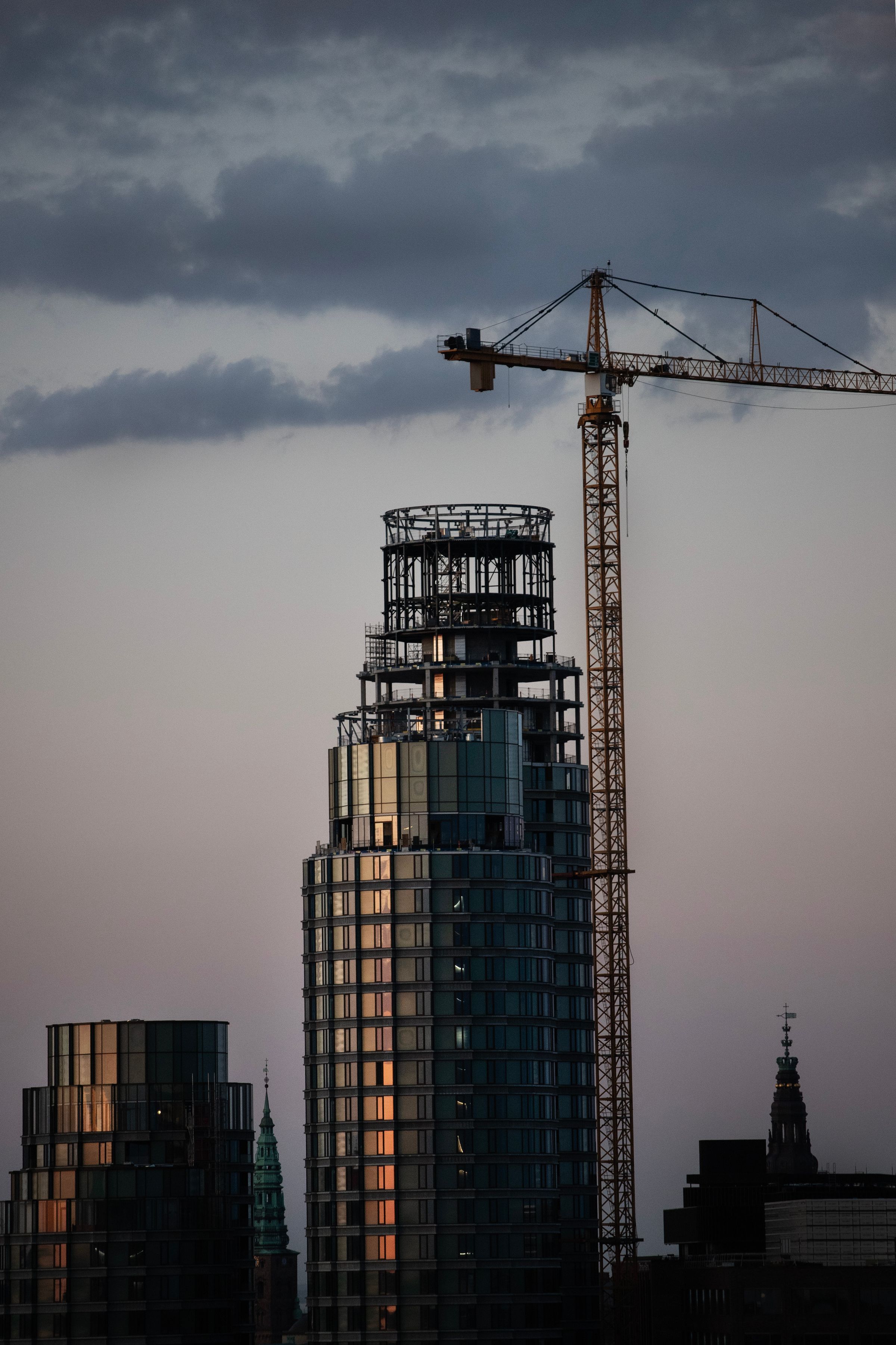 A tower under construction with a crane, against a grey dusk sky
